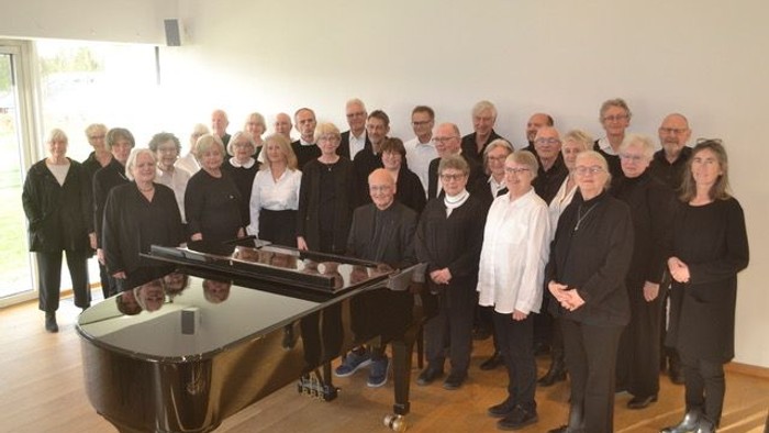 En gruppe af mænd og kvinder står sammen med et piano i en rummelig lokalitet