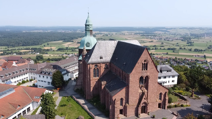 Ein großer Ziegelbau mit hohem Turm, umgeben von Gebäuden und Grünflächen, in ländlicher Umgebung.