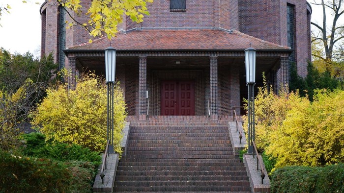Breite Treppe, flankiert von hohen Laternen, führt zu einem Backsteingebäude mit roter Doppeltür.