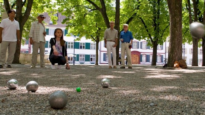 Eine Gruppe von Menschen spielt Boccia auf einem Kiesplatz unter Bäumen.