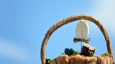 Ein Weidenkorb hält ein Ei und kleine Pflanzen, vor einem klaren blauen Himmel.