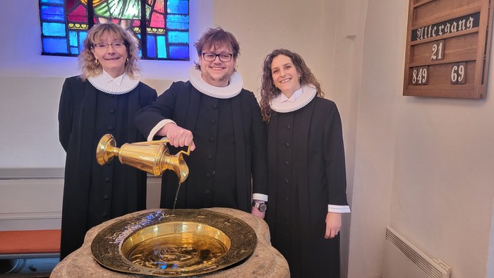 Tre personer i ceremonielle kjortler, der udfører en dåbsritual med vievand i en kirkelig ramme.