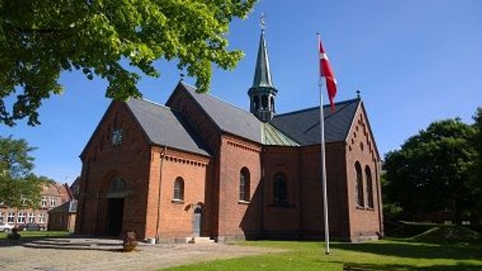En rød murstenskirke med spiret og en flagstang med flag.