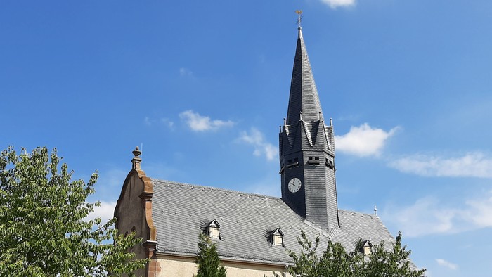 Kirche mit hohem Turm und Uhr, umgeben von Bäumen und Blumen.