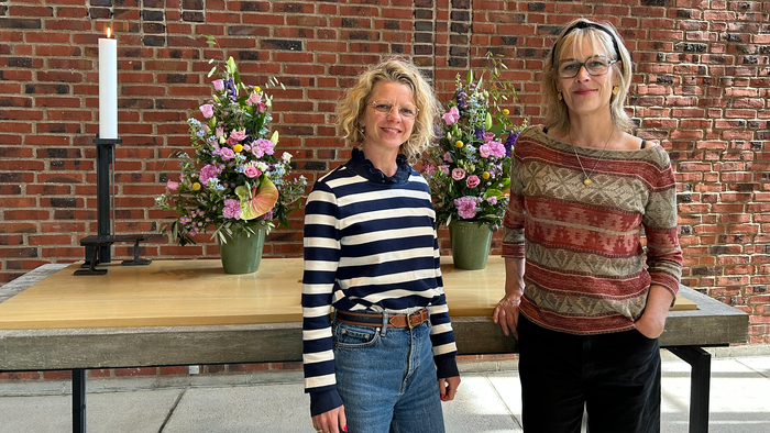 To kvinder står ved siden af et træbord med blomsterdekorationer og et stearinlys i et rum med murstensvægge.