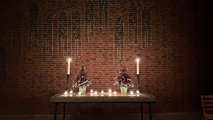 Mindesalter med tændte lys og blomsterarrangementer foran en murstensvæg med mindetavler.