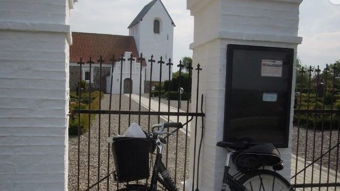 En svart cykel er låst til en sort stang i en hvid mur med en sort dør og en kirke i baggrunden