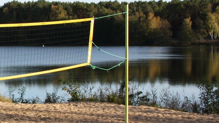 Volleyballnetz am sandigen Strand in der Nähe eines ruhigen Sees mit Bäumen im Hintergrund.
