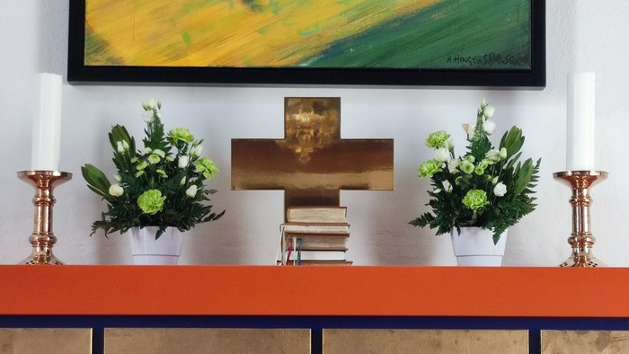 En håndværkerkrucifix på en rød og guld farvet alter med to blomsterarrangementer og to lysekander.