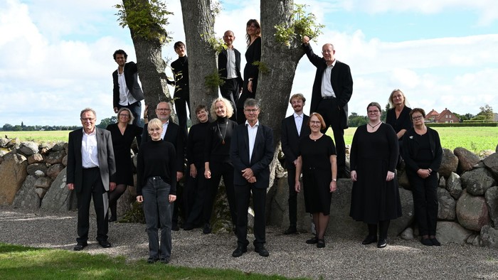 Gruppe af mennesker i formelt tøj, der poser sammen udendørs nær en stenmur og træer.