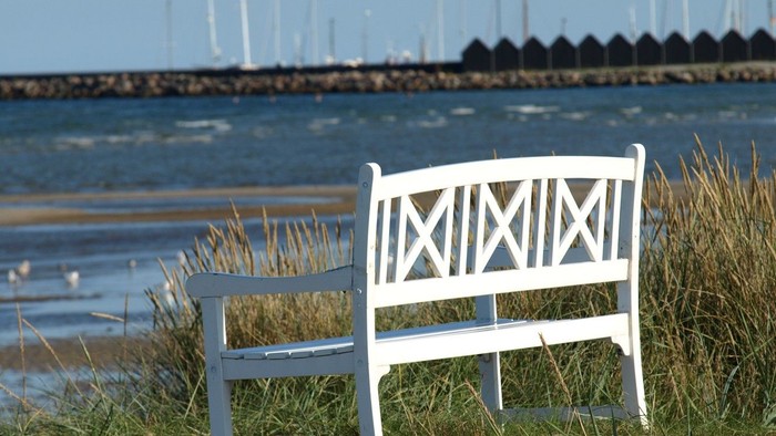 En fredfyldt hvid bænk med udsigt over et roligt vand med sejlbåde og en fjern marina.