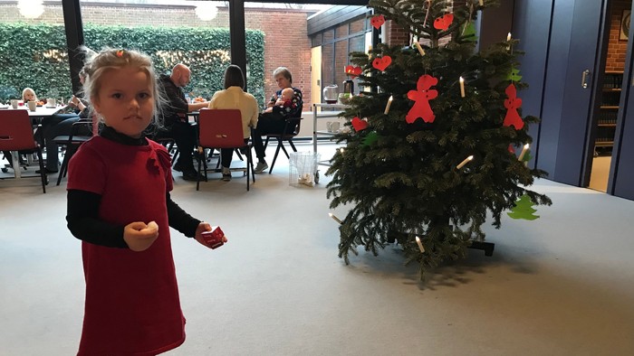 En ung pige i en rød kjole står ved siden af et pyntet juletræ i et rum, hvor der sidder mennesker ved borde.