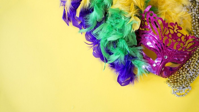 Farverig Mardi Gras-maske pyntet med farverige fjer og perler på en lys gul baggrund.