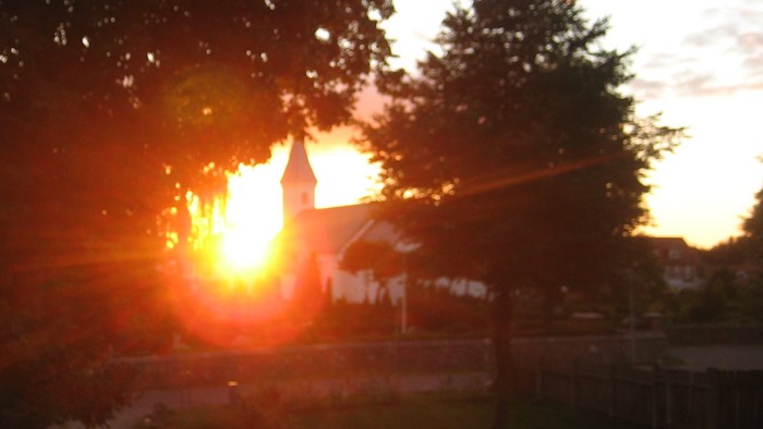 Solnedgang set over en vej med træer og en kirke i baggrunden