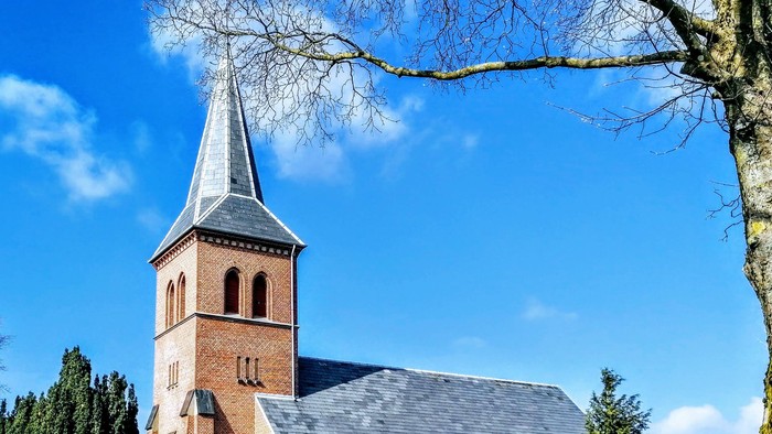Murstenskirke med højt tårnspir og buede vinduer, omgivet af træer og grønne omgivelser under blå himmel.