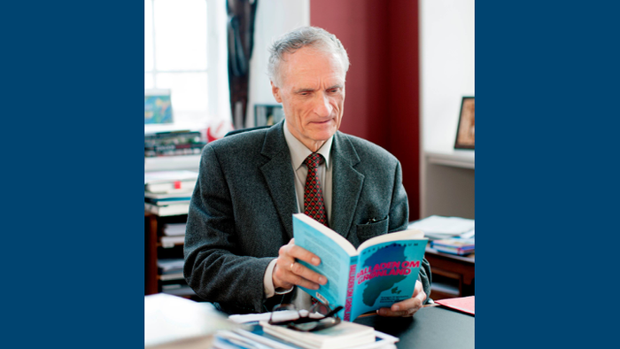 En ældre mand i jakkesæt læser en åben bog ved et skrivebord på et kontor.