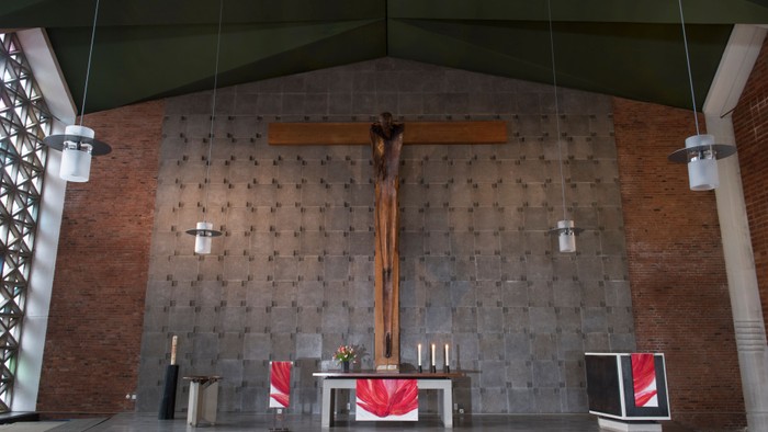 Die Kirche zeigt einen Altar mit Kreuz und Kerzen, flankiert von zwei roten Teppichen und einem Bildschirm.