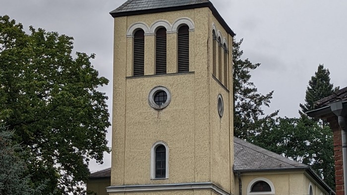 Kirchturm mit Kreuz und Uhr, umgeben von Bäumen und Büschen