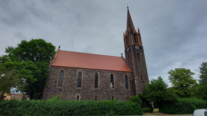 Ein Kirchengebäude mit hohem Turm und rotem Dach, umgeben von Bäumen und Büschen.