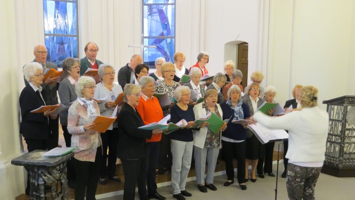 Ein gemischter Chor probt in einem hellen Raum mit großen Fenstern.