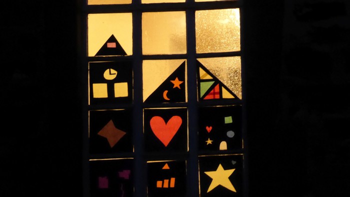 Ein Fenster mit bunten geometrischen und herzförmigen Ausschnitten, das nachts von hinten beleuchtet wird.