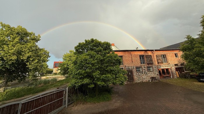 Regenbogen über einem Backsteingebäude mit mehreren Fenstern, umgeben von Bäumen und einem Zaun.