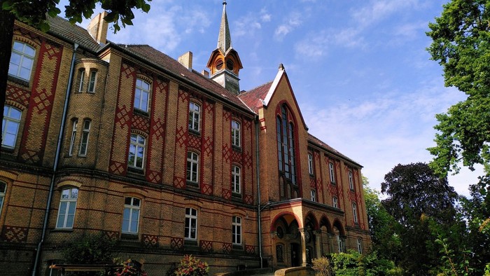 Historisches Backsteingebäude mit spitzem Turm und großen Fenstern, umgeben von Bäumen