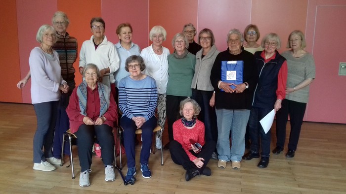 Gruppe von Senioren, die für ein Foto posieren, einige sitzend und einige stehend, in einem Raum mit orangenfarbenen Wänden.