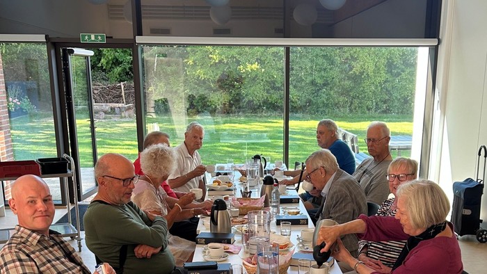 En gruppe ældre personer sidder ved et langt bord med mad og drikkevarer i et lys og moderne rum med udsigt til en have.