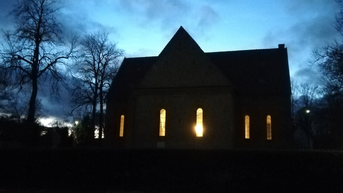 En mørk kirke med lysende vinduer og træer i forgrunden under et blåtintet nattehimmel.