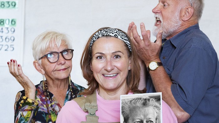 Tre personer poser glædeligt, mens de holder et indrammet portræt og vinker entusiastisk foran et nummereret skilt.