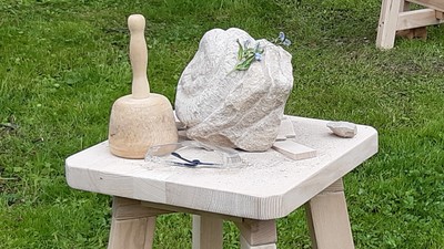 Auf einem Holztisch im Freien stehen ein großer Stein, ein Hammer und andere Werkzeuge.