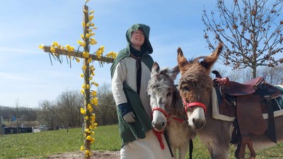 En kvinde i grøn kostume står ved et kors med gule blomster og holder en hest.