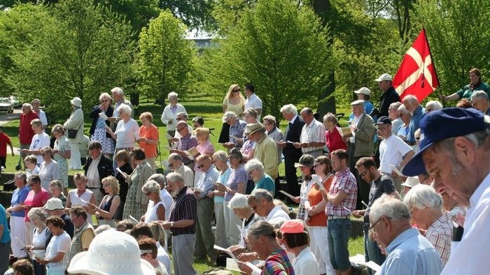 En folkemængde er samlet i en park, hvor en rød flag med hvid krone er opstillet.