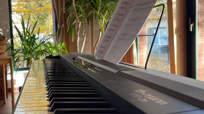 Klaviatur eines Klaviers mit Notenblatt im Notenständer, in der Nähe eines Fensters mit Pflanzen.