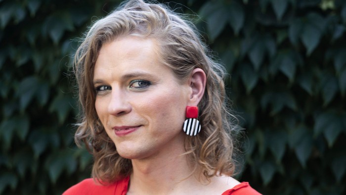 Frau in rotem Hemd mit Ohrringen, die vor grünem Blattwerk steht.