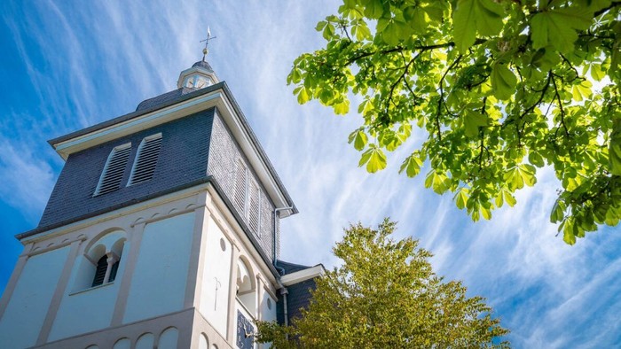 Ein historischer Kirchturm, eingerahmt von üppigem grünem Laub vor einem klaren blauen Himmel.