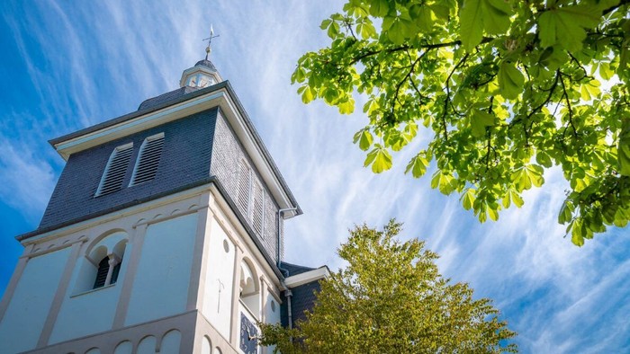 Ein historischer Kirchturm, eingerahmt von üppigem grünem Laub vor einem klaren blauen Himmel.