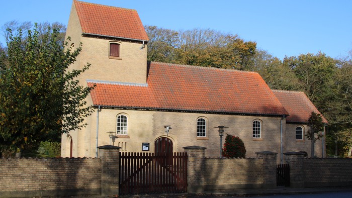 Et lille kirke med røde tag og murstensmure, omgivet af træer.