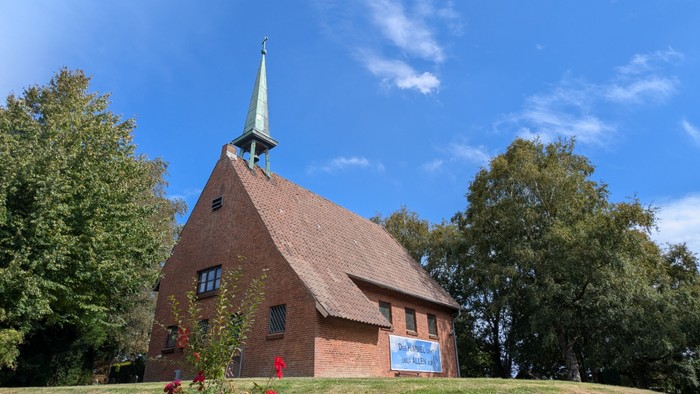 Die Kapelle Westerthal im Sommer. Der Himmel ist blau mit einigen kleinen Wolken. Um die Kirche stehen Laubbäume, im Vordergrund ragt ein Busch ins Bild.