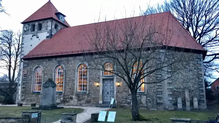 Alte Kirche mit rotem Dach und Turm, umgeben von Bäumen und Gräbern