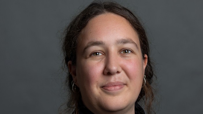 En kvinde med mørkt skulderlangt hår og store øreringe, iført en mørk top, smiler blidt mod en grå baggrund.