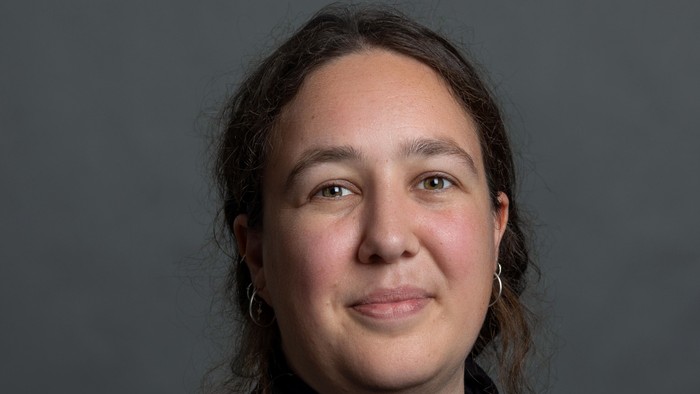 En kvinde med mørkt skulderlangt hår og store øreringe, iført en mørk top, smiler blidt mod en grå baggrund.