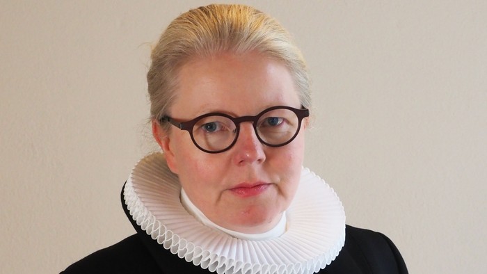 En kvinde med blond hår og briller bærer en sort jakke med hvid krage.