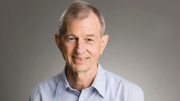 En smilende ældre mand iført en lyseblå skjorte med krave på en ensfarvet baggrund.