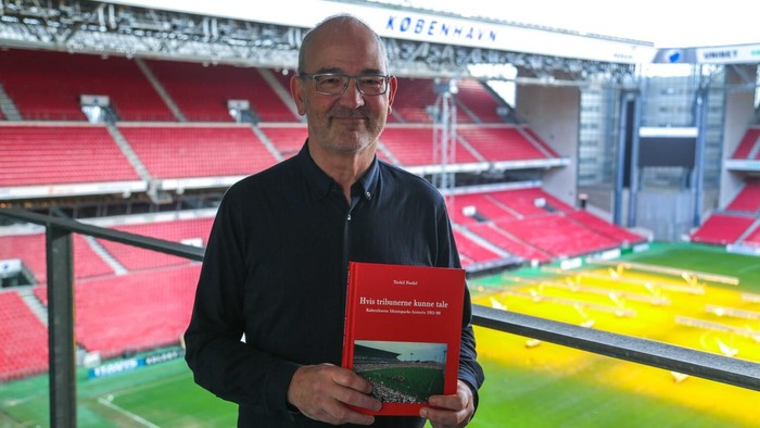 En mand holder en bog på et stadion, stående nær en tom rød sidesektion.