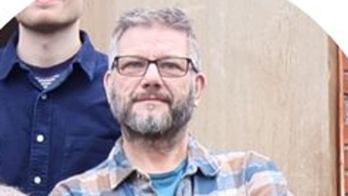 En gruppe mennesker står udendørs med en mand i enternet skjorte, der poserer selvsikkert forrest.