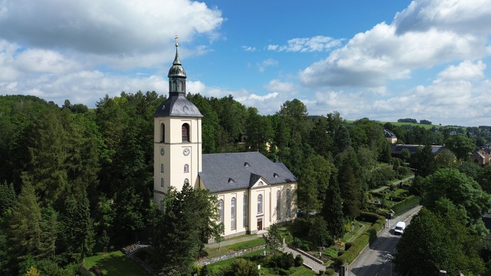 Ein weißer Kirchturm mit Uhr und Kreuz steht inmitten von Bäumen und Gebäuden in einer grünen Landschaft.