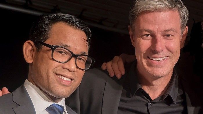 Zwei lächelnde Männer in Anzügen, einer mit Brille.