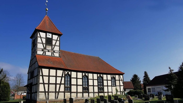 Bunte Kirche mit hohem Turm und Uhr, umgeben von Friedhof und Bäumen unter klarem Himmel